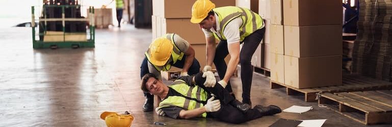 Industrial worker injured in a warehouse accident, while co-workers try to help him.