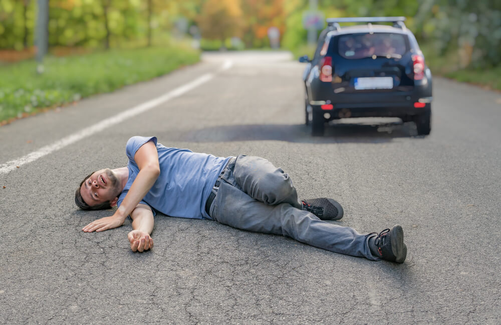 Injured man on road after hit-and-run accident in Greensboro, NC, with fleeing vehicle in background.